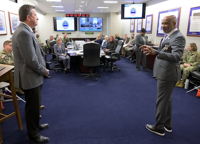 Two men face one another in a conference room with men and women sitting at a table in the background.