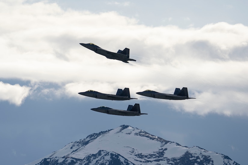 Four military jets fly in formation with one further away from the others and pointing upwards. A snow-covered mountain is in the background.