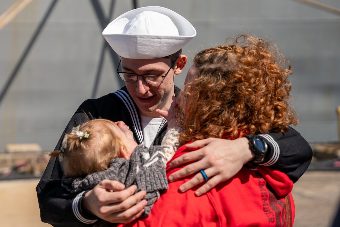 A sailor hugs a person holding a baby. The sailor looks down at the baby and smiles.