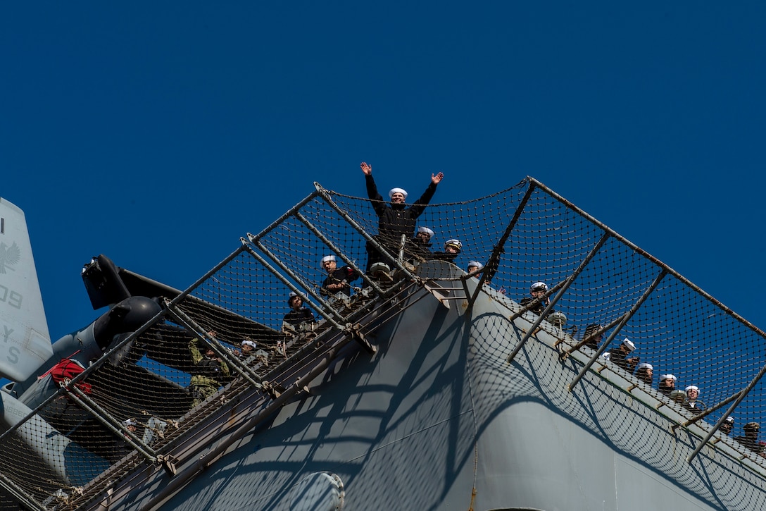 Soldiers look down from the flight deck of a Navy ship as one raises their hands in excitement.