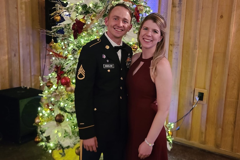 A uniformed service member and person in an evening dress pose in front of a Christmas tree.