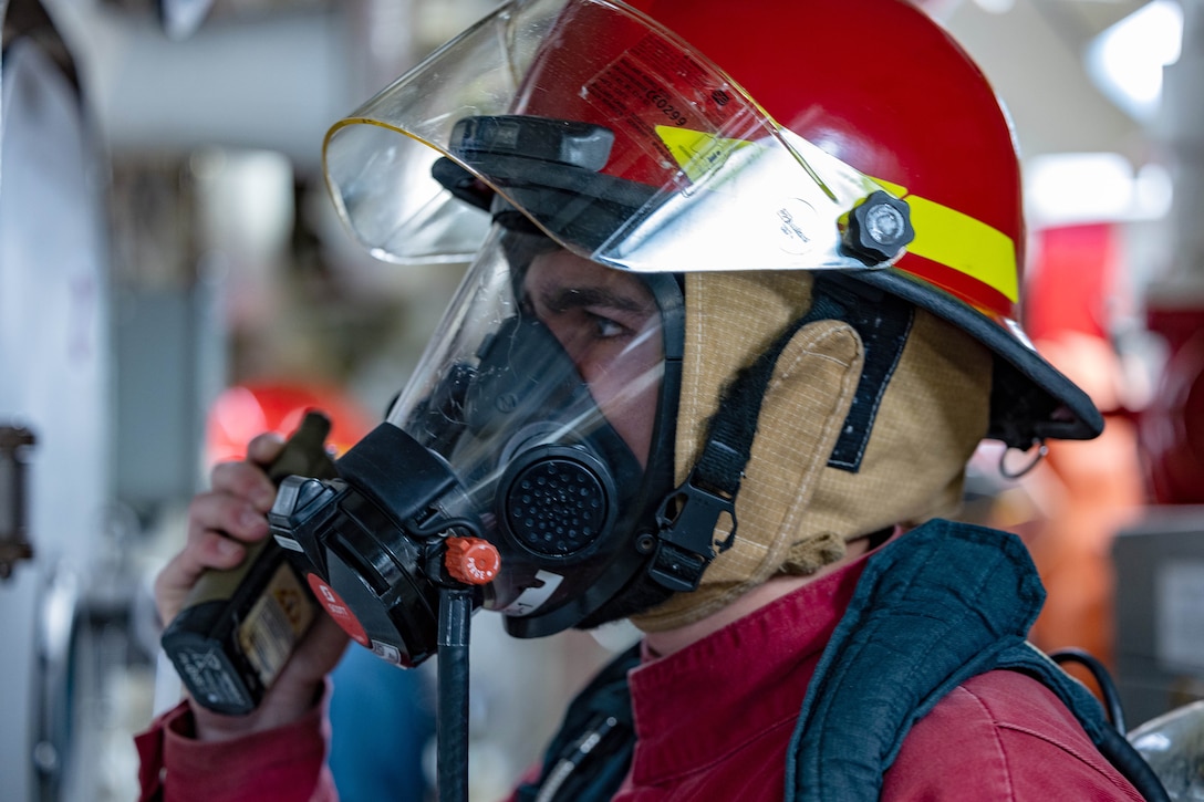 A side view of a sailor in firefighting gear talking on a walkie talkie in a maintenance room.