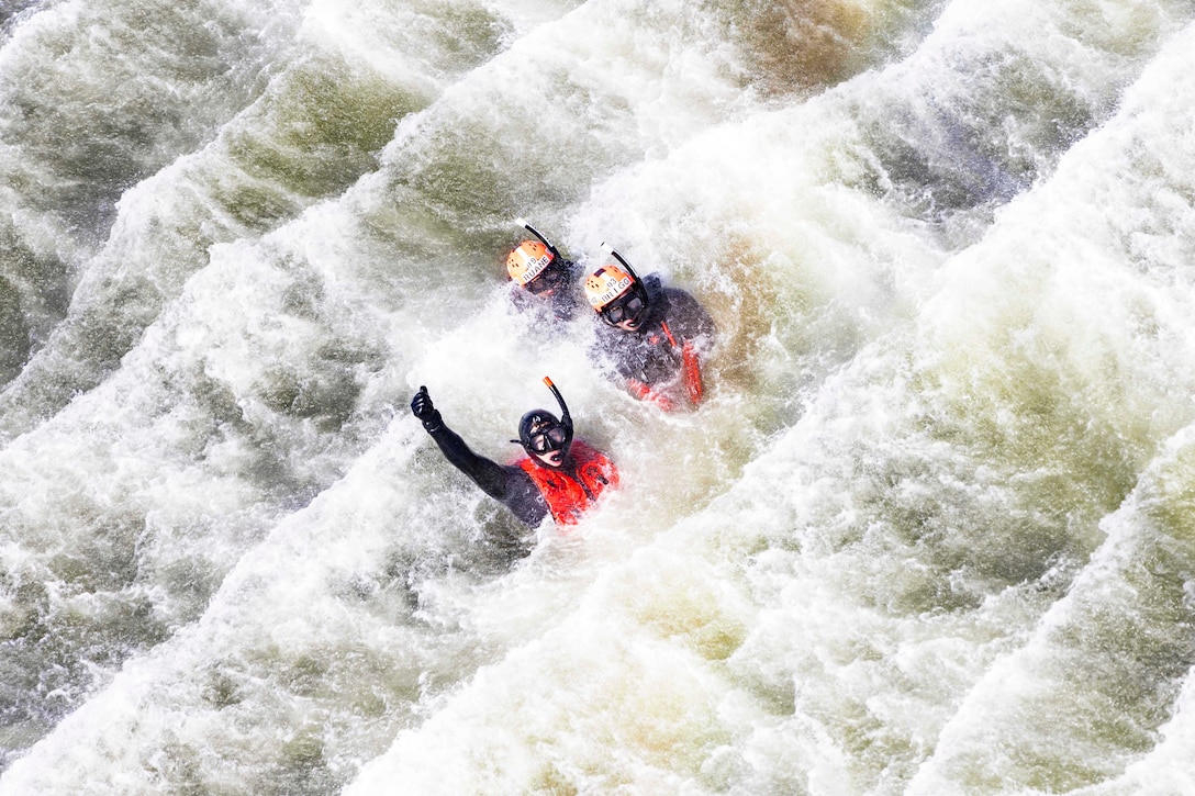 One of three sailors raises their hands while treading in rough waters as seen from above.