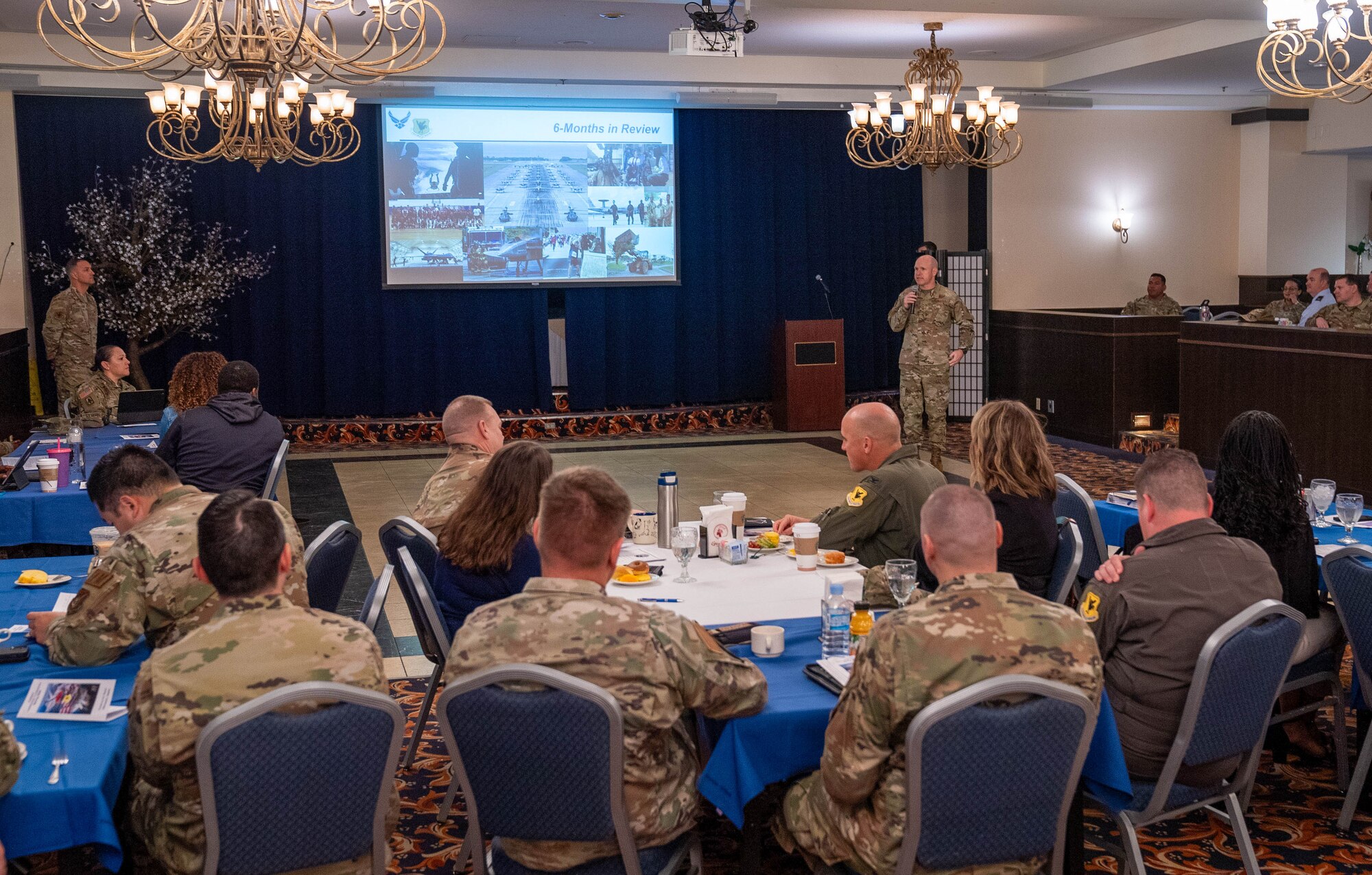 Airmen sit at tables listening to the speaker at the front of the room
