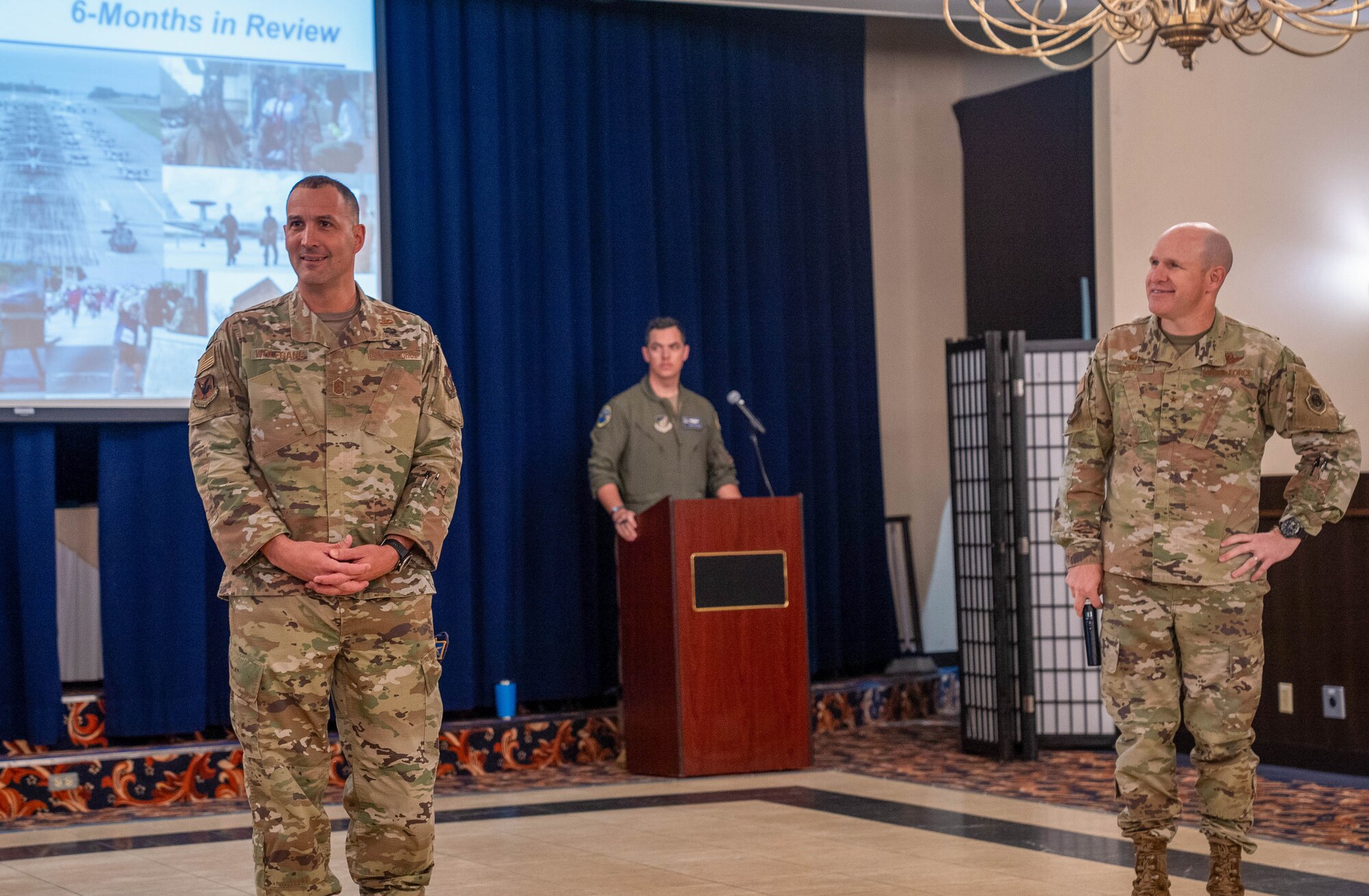 Three Airmen stand at the front of a room in front of a audience
