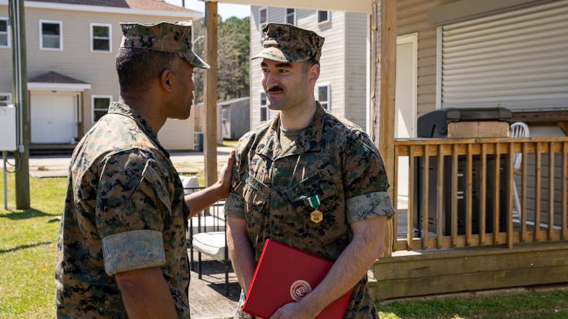 A Marine holds an award while another puts a hand on the Marine with houses in the backdrop.