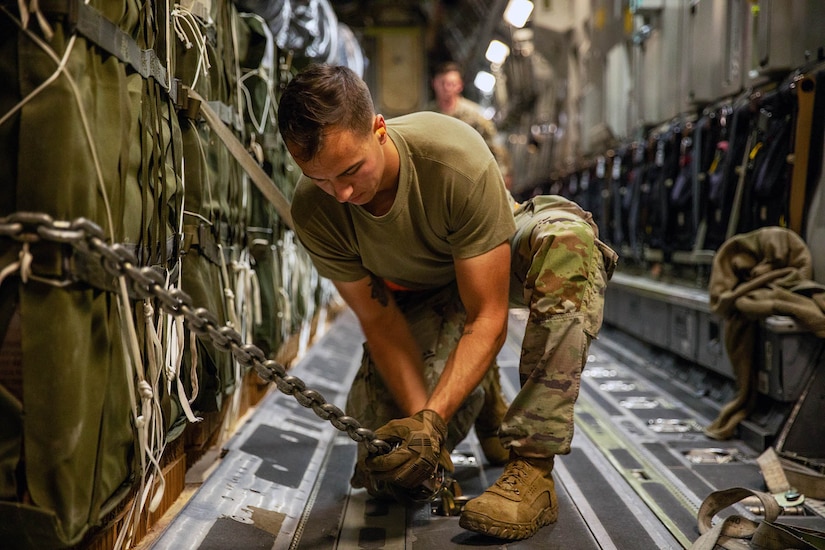 A kneeling airman secures a chain that is wrapped around cargo to the floor of an aircraft.