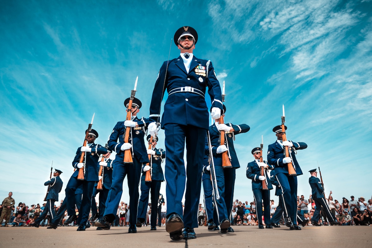 Airmen in formal uniforms and holding rifles march toward the camera and in other directions. A crowd of people watches.