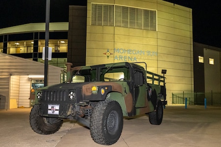 A military vehicle painted in dark camouflage is parked in front of a large arena. The arena's sign reads "Mohegan Sun Arena".