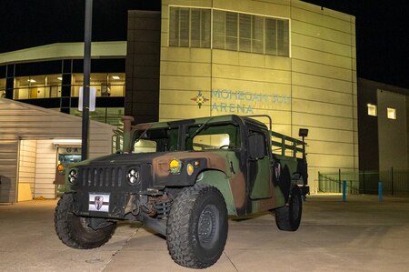 A military vehicle painted in dark camouflage is parked in front of a large arena. The arena's sign reads "Mohegan Sun Arena".