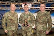 Three U.S.Army Servicemembers pose for a photo, the center Servicemen holds an award. Behind them is an empty ice hockey arena.