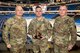 Three U.S.Army Servicemembers pose for a photo, the center Servicemen holds an award. Behind them is an empty ice hockey arena.