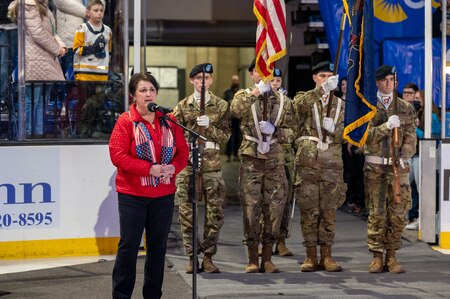 A woman stands at a microphone while singing. Behind her are U.S.Army Servicemembers.