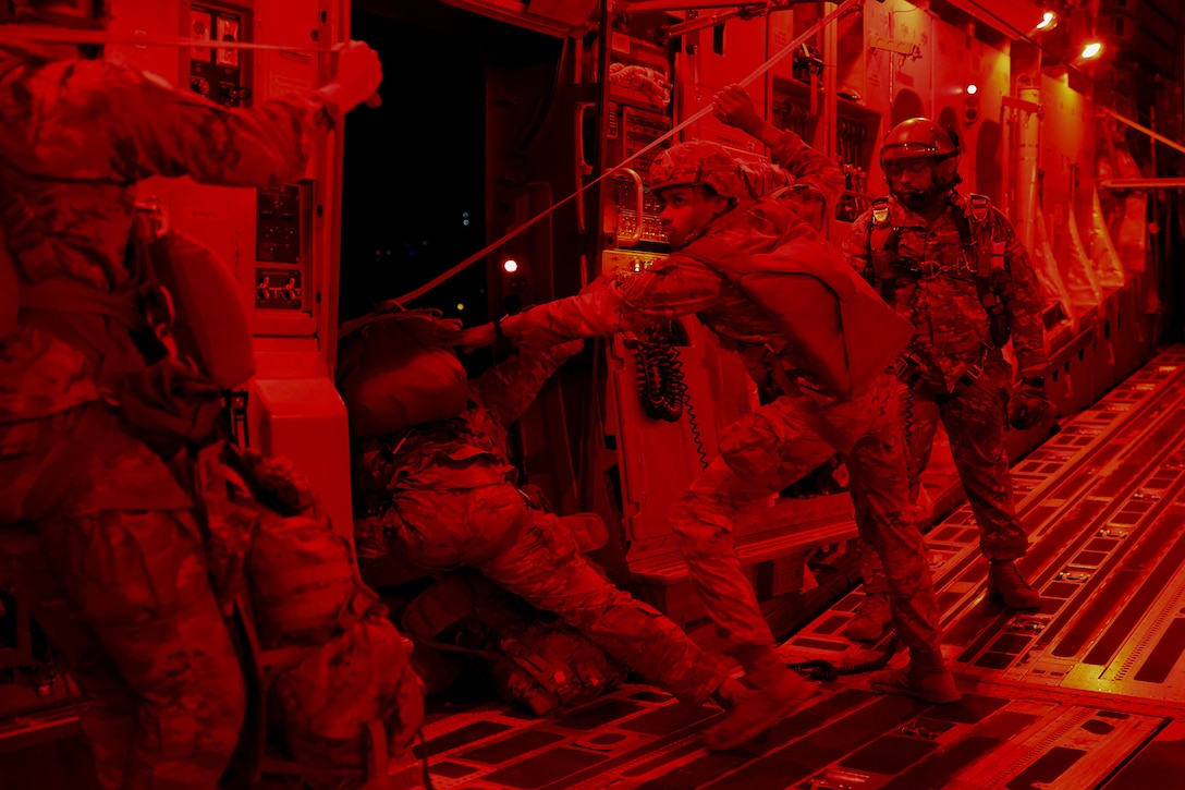 An Army paratrooper leans out the side of a military aircraft as others stand inside during nighttime training. The aircraft is illuminated by a red light.