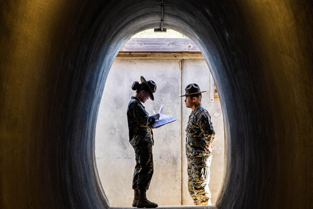 Two Marines stand as one looks down at a clipboard. They are photographed through a tunnel like structure.