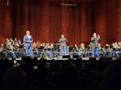 An Army soldier in a dark ceremonial uniform is holding a microphone in one hand and gesturing with the other while singing on a stage in front of other Army Soldiers in similar uniforms who are performing on various musical instruments.  There is another man on stage who is wearing a blue suit with no tie and is looking towards the person who is signing.