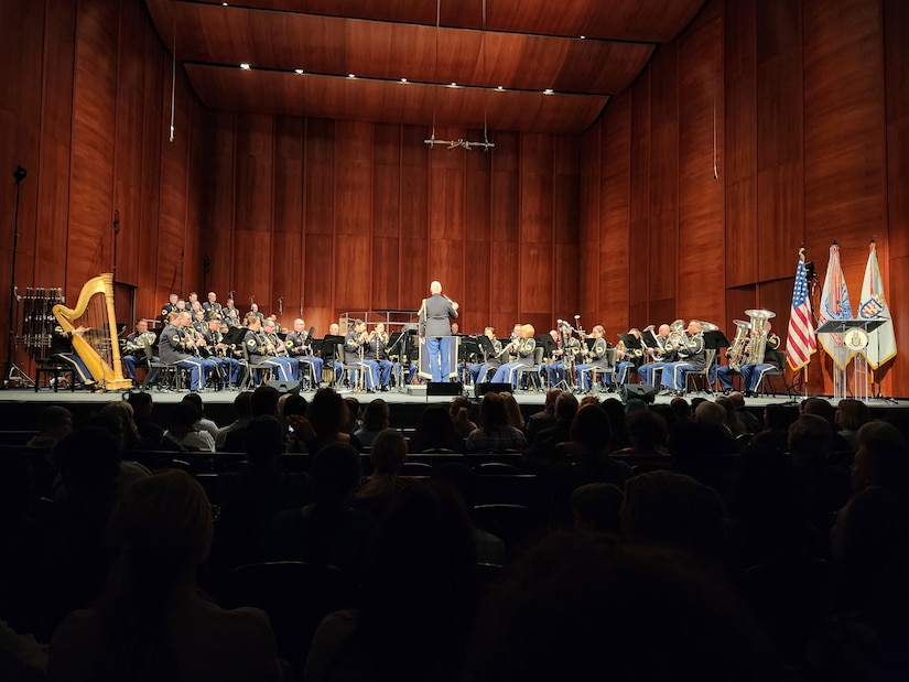 A military band dressed in dark ceremonial uniforms is performing on a stage with a conductor in the middle.