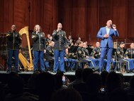 Three people dressed in dark ceremonial Army uniforms are holding microphones and singing on a stage next to a man in a blue suit, no tie, who is also singing into a microphone. There is an Army band in dark ceremonial unifoms seated and performing in the background.