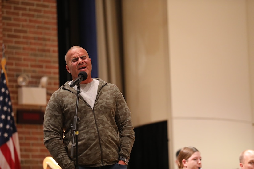 A man dressed in blue jeans and a grey cardigan sweater is singing with his eyes closed in front of a microphone that is on a mic stand.