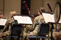Army soldiers wearing green camouflage uniforms are seated facing away from the camera while performing on musical instruments on a stage.