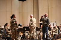 A man dressed in blue jeans and a grey cardigan sweater is holding a microphone and singing on stage towards a woman wearing an Army green camouflage uniform and holding a microphone. In the background are musicans wearing Army green camouflage and performing on musical instruments, while a man in the center is conducting.