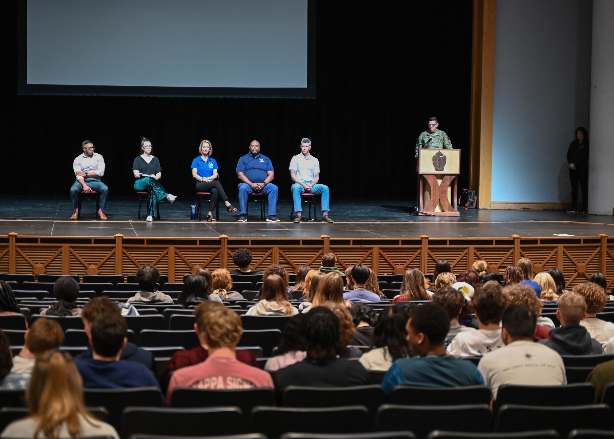 Panel members sit on stage as man addresses high school students.