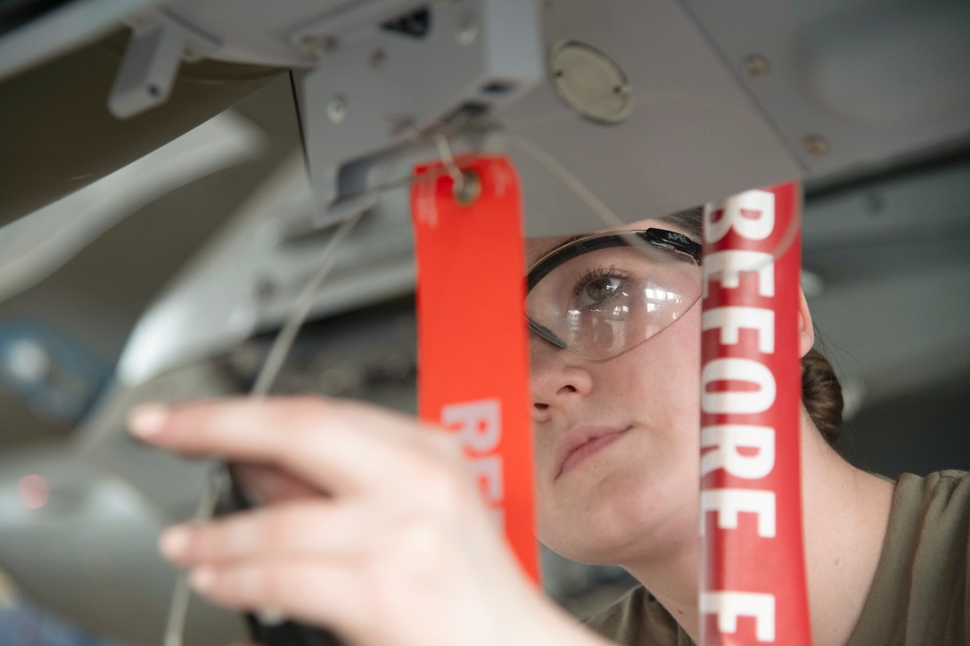 An airman wearing safety glasses holds a wire while working under an aircraft.