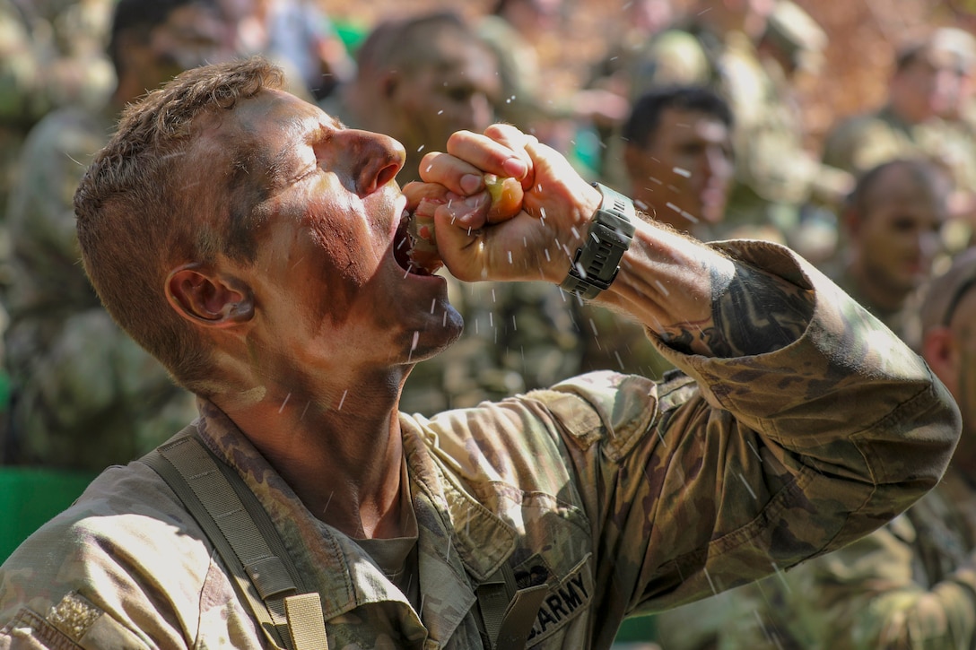 An apple splashes juice as a soldier wearing camouflage squeezes it into their mouth as fellow soldiers sit in the blurred background.