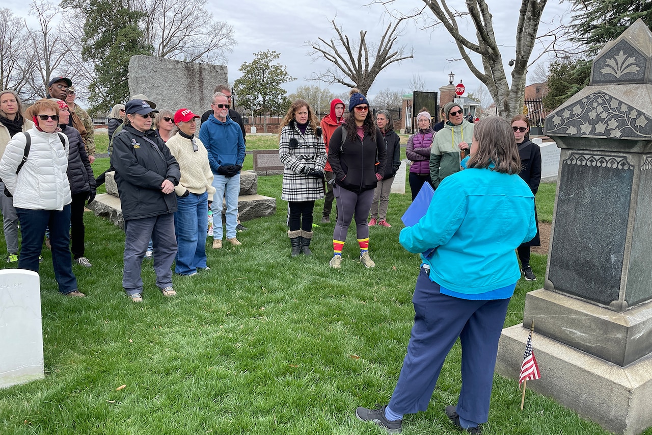 A person standing beside a grave faces several other people who are watching as the person speaks.