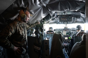 A military member stands in the cockpit of an aircraft and watches two pilots fly the aircraft.
