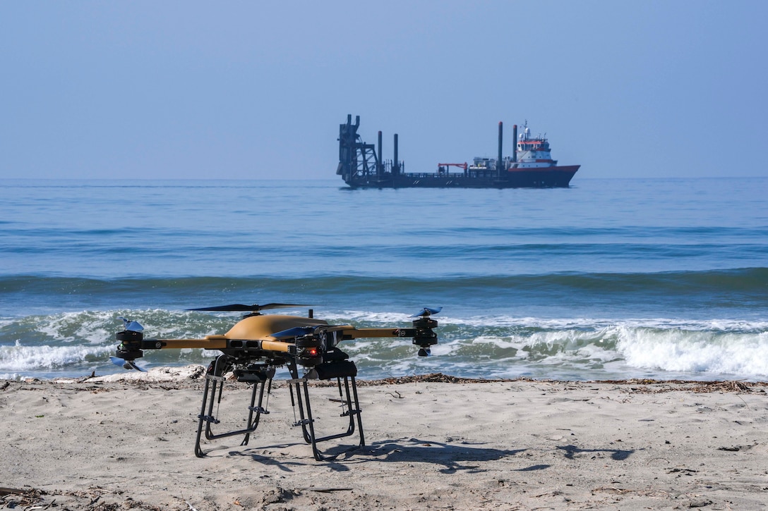 A yellow drone is on a beach with waves lapping at the shore and a ship in the distance.