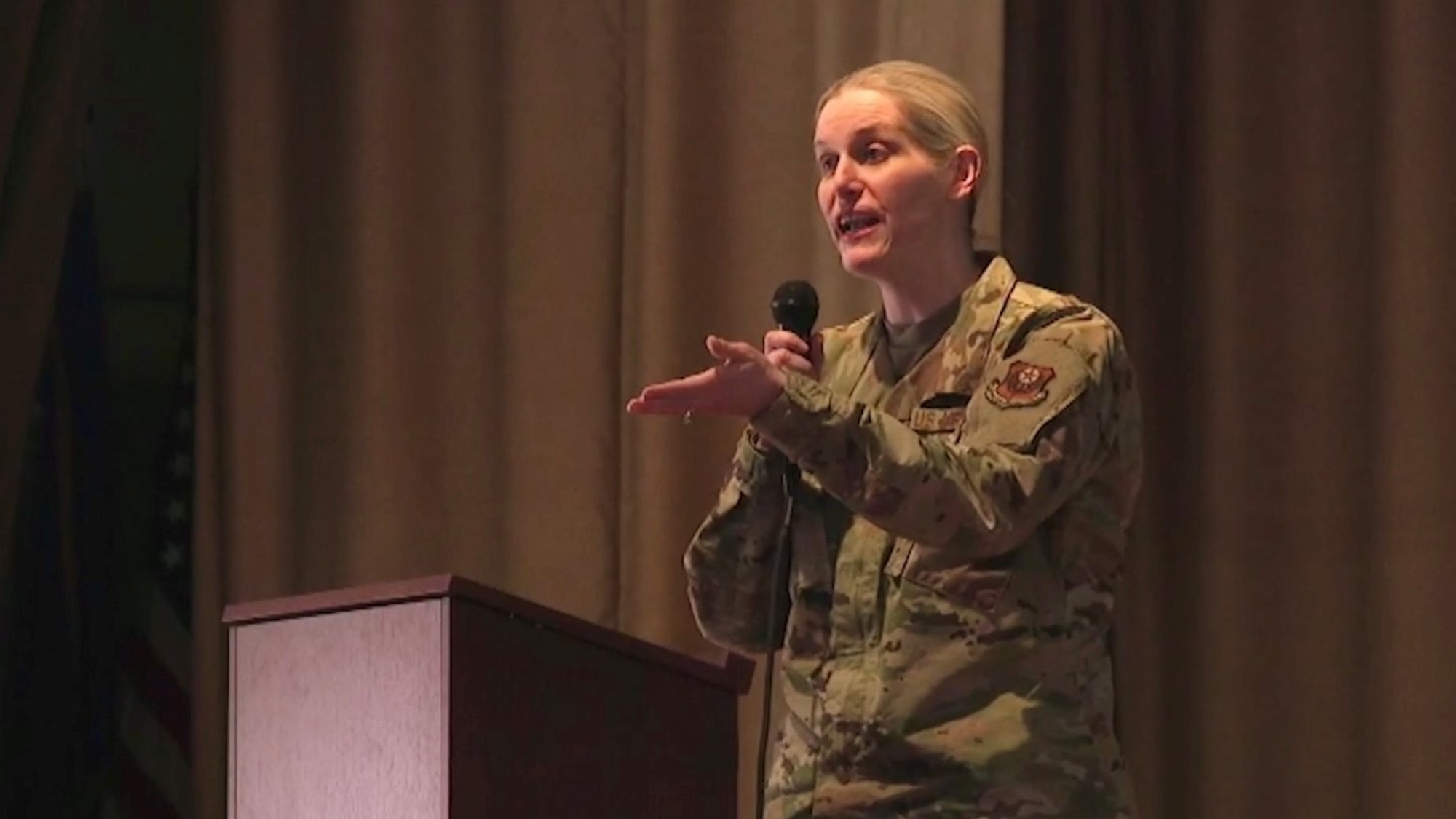 A woman in military uniform stands behind a podium on stage speaking into a microphone she is holding.