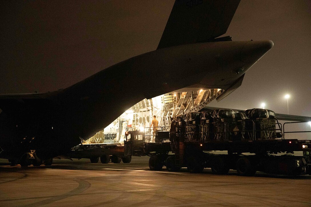 Personnel  work with cargo on a platform next to the lighted back opening of a parked aircraft against a dark sky.