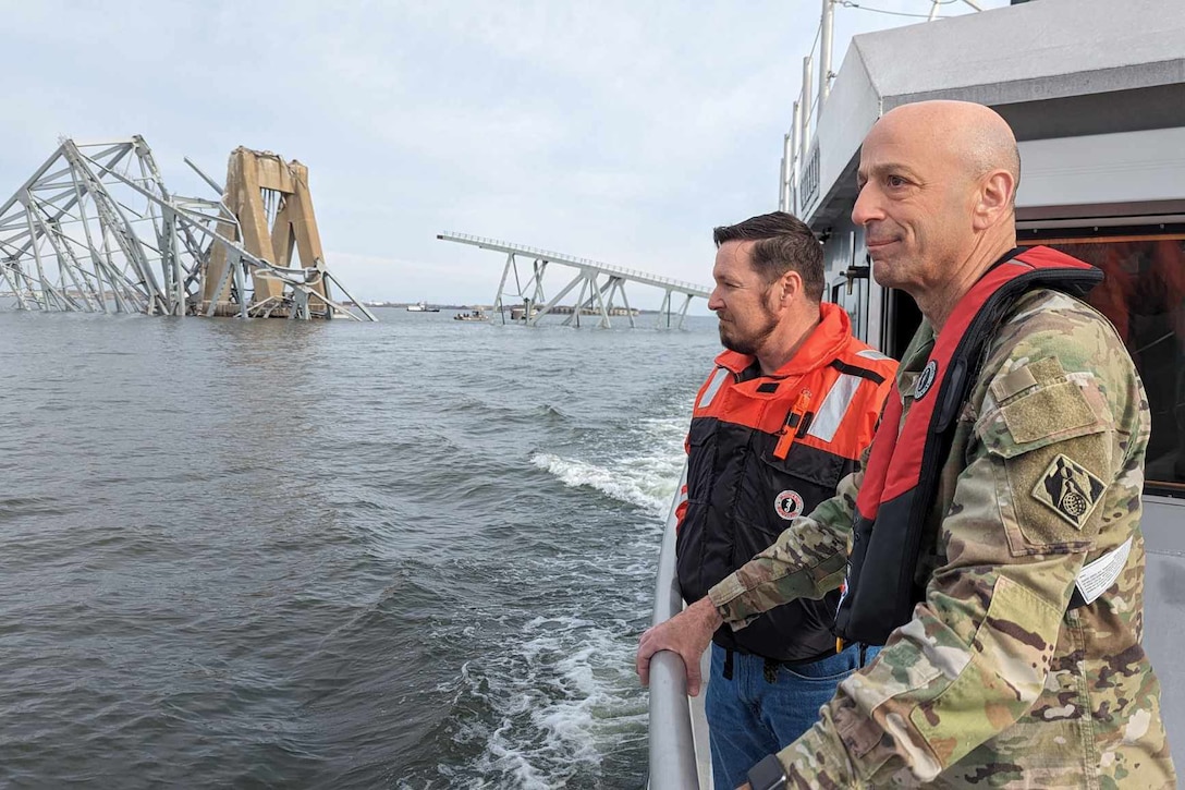 A military officer and a civilian stand on the side of a boat and look at bridge that collapsed in the water.