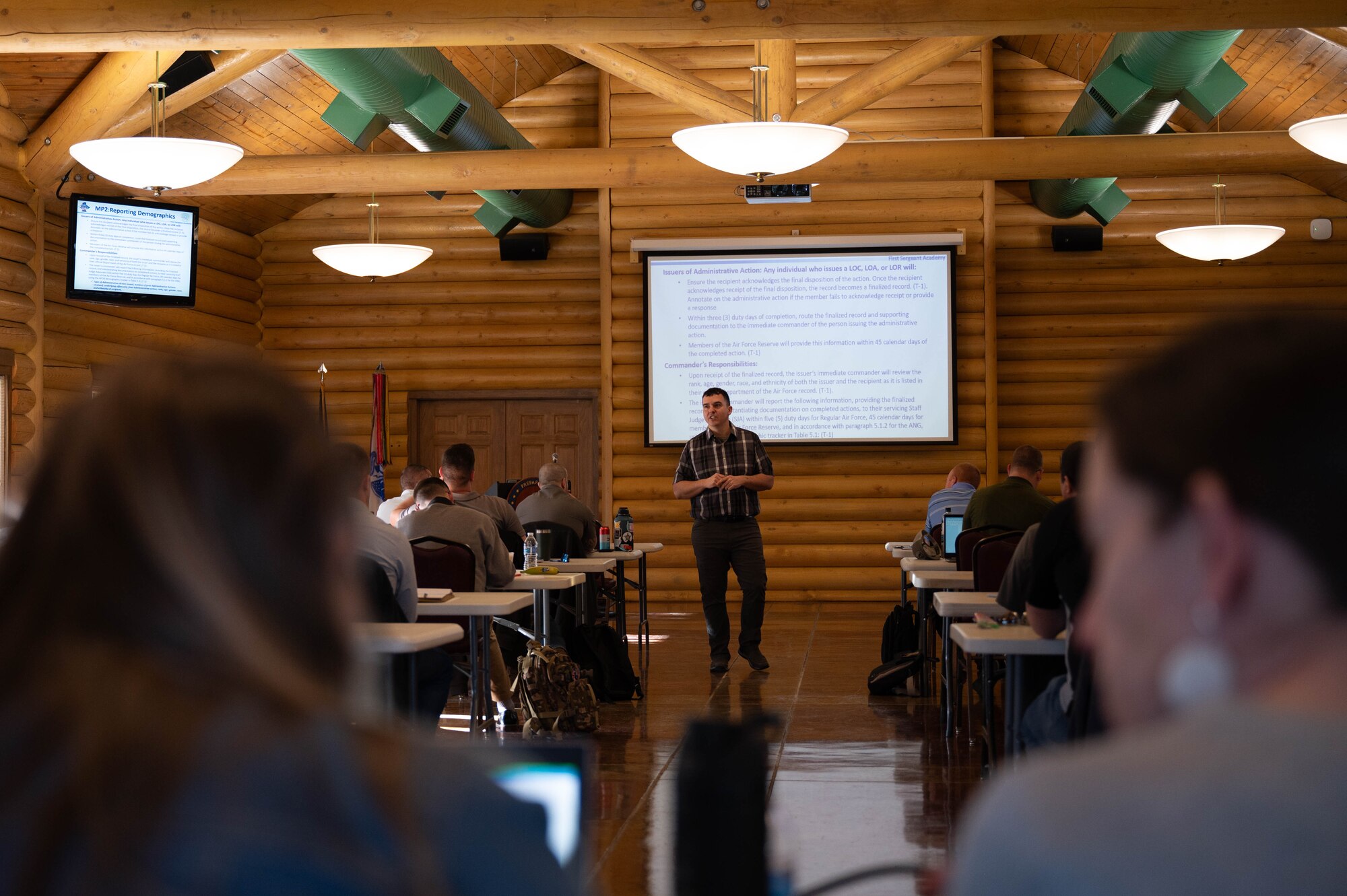 A man, standing in front of a projector screen, speaks to a crowd.