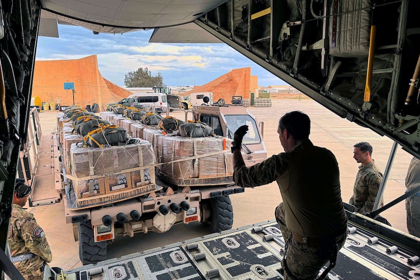 An airman, shown from behind, kneels inside the open back of aircraft and waves toward a truck backing up with cargo.
