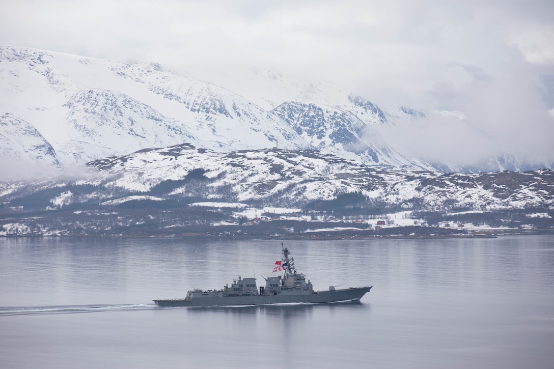 A large military ship steams through the water with snowy mountains and clouds in the background.