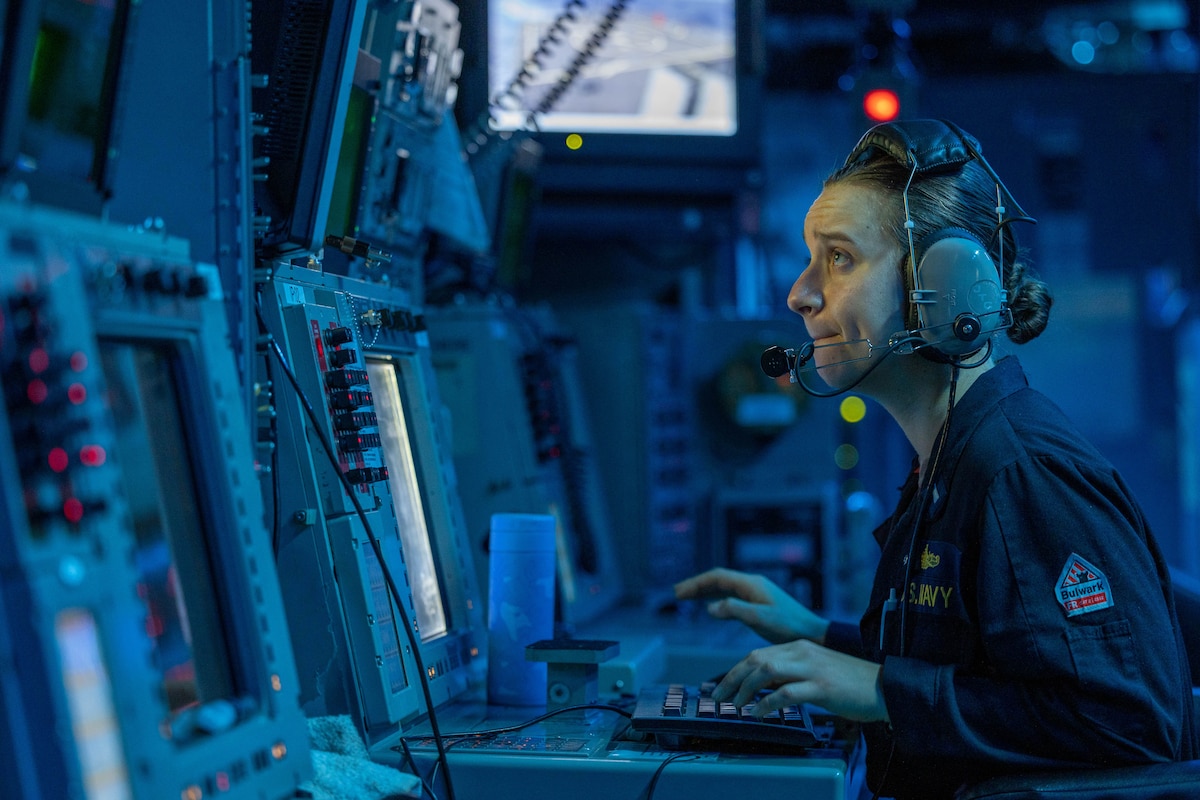 A sailor wearing a headset sits in front of an array of monitors and controls in a darkened blue-lit room.
