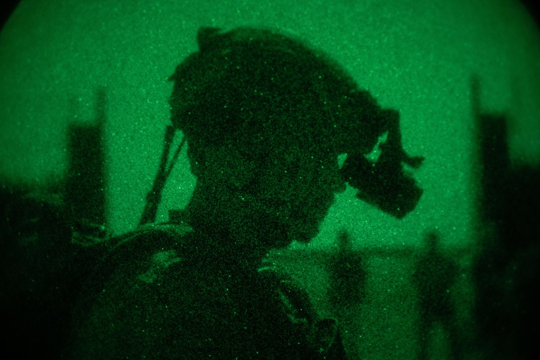 A nighttime close-up of a soldier wearing a helmet and looking down. The photo is illuminated by a green light.