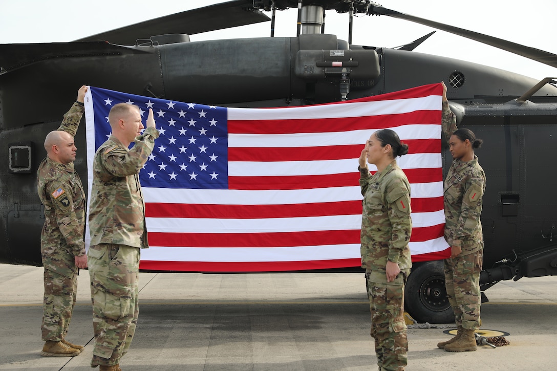Two soldiers raise their right hands while facing one another as two soldiers hold the American flag in the background. A military aircraft is parked on the tarmac.