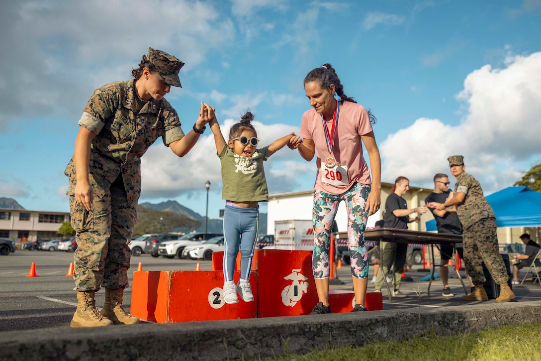 A Marine and a participant help a child down from a podium with a parking lot in the background.