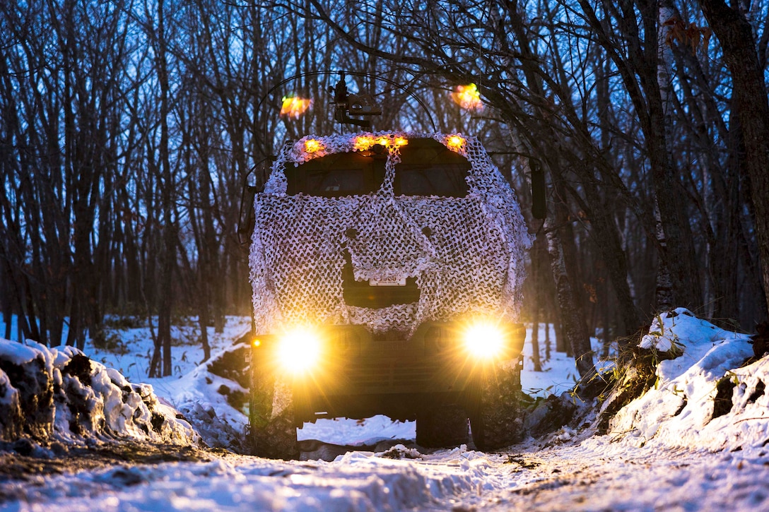 An armored vehicle covered in netting moves through the snowy woods illuminated by headlights and top lights.