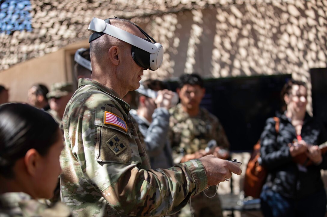 A soldier wears a virtual reality headset with controllers in hand while other soldiers watch the demonstration.