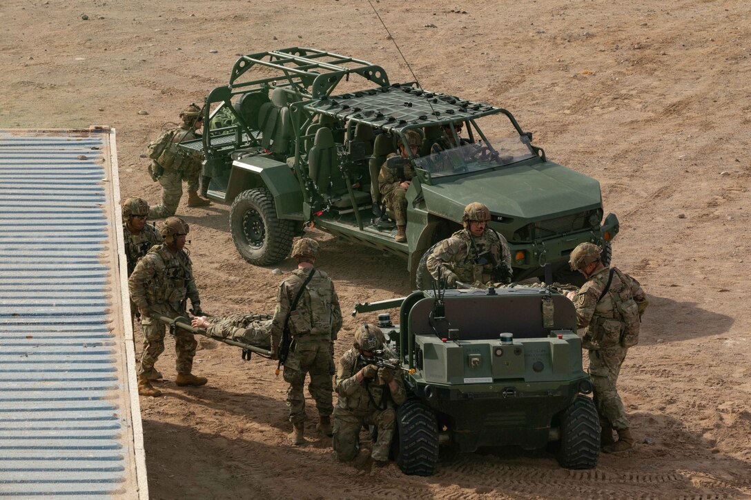 Soldiers carry a simulated patient on a gurney while others operate a small and large vehicle in a desert area with a metal roof cover to the side.