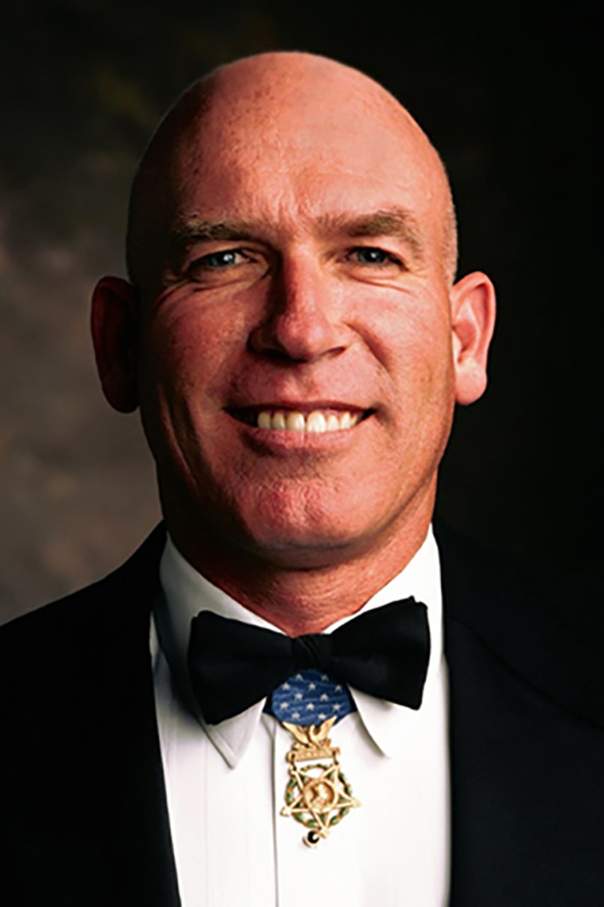 A person wearing a tuxedo and a medal around their neck smiles for a photo.