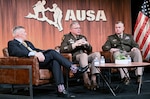 Three men sit in chairs on a stage in front of a wall with a sign that says AUSA.