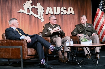 Three men sit in chairs on a stage in front of a wall with a sign that says AUSA.