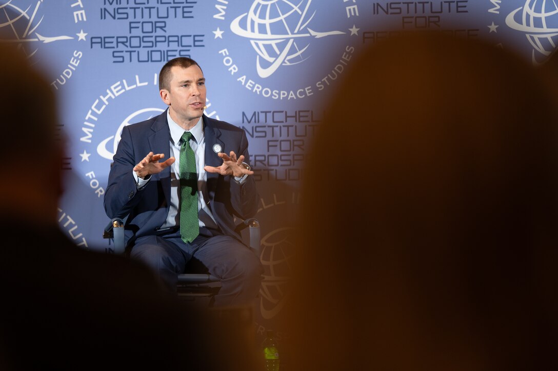 A man sits in a chair. Behind him, a backdrop has earth-shaped logos and words that says, "Mitchell Institute for Aerospace Studies."