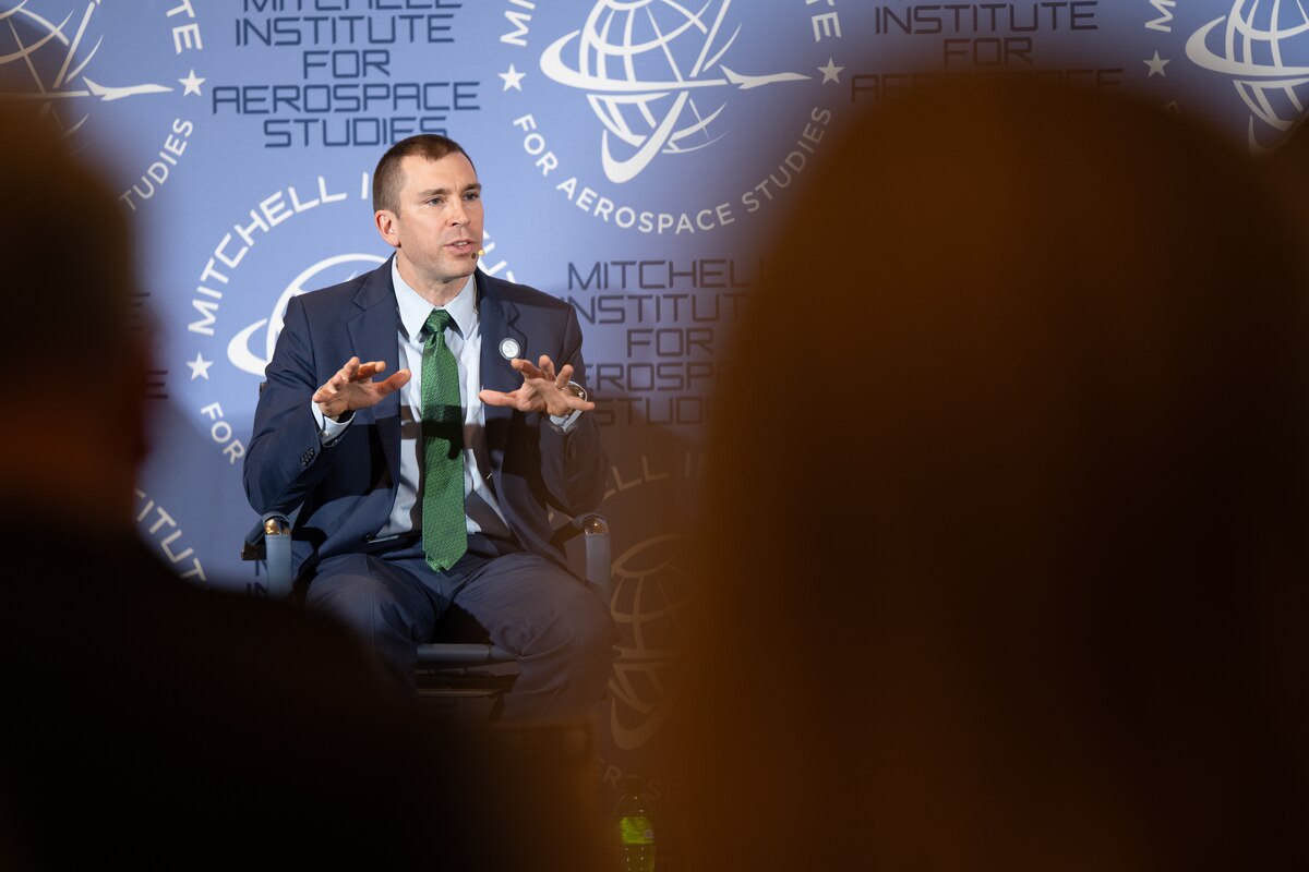 A man sits in a chair. Behind him, a backdrop has earth-shaped logos and words that says, "Mitchell Institute for Aerospace Studies."
