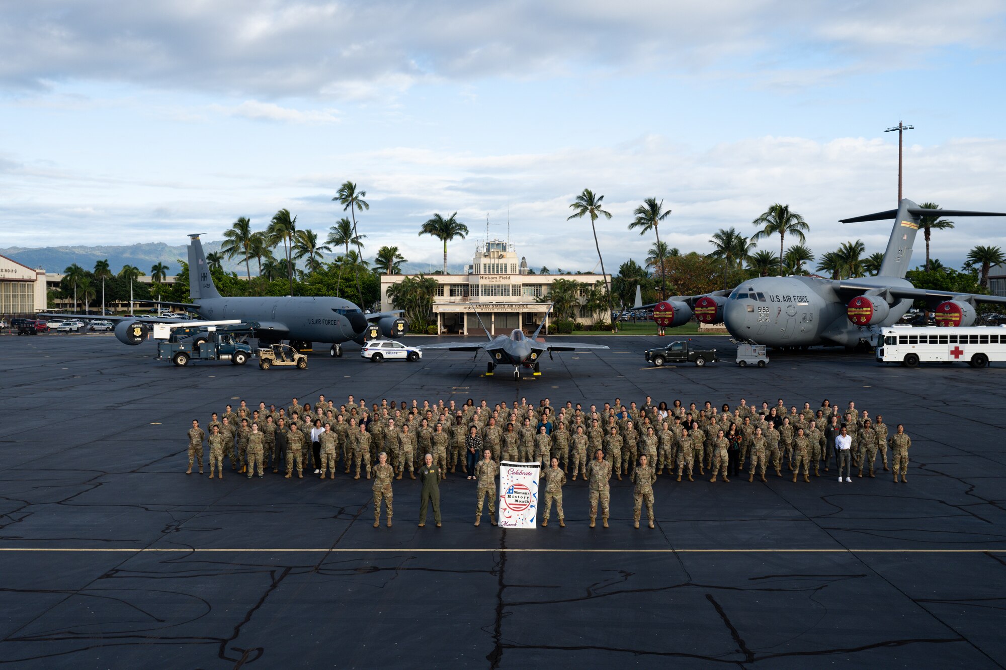 Women assigned to Joint Base Pearl Harbor Hickam stand for a group photo with static aircraft in the background